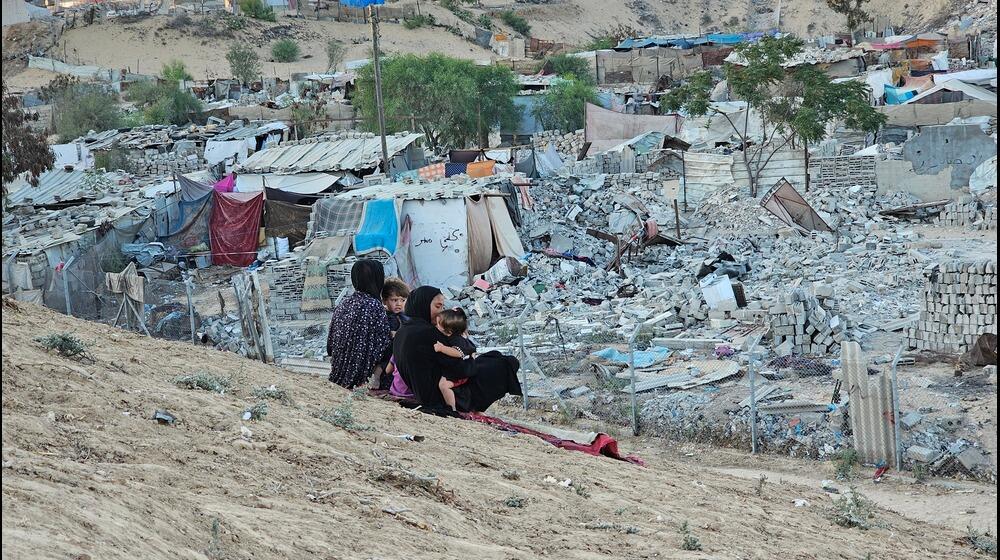 Two women, each holding a baby, sit next to each other on the side of a small mound, overlooking the rubble in Gaza.